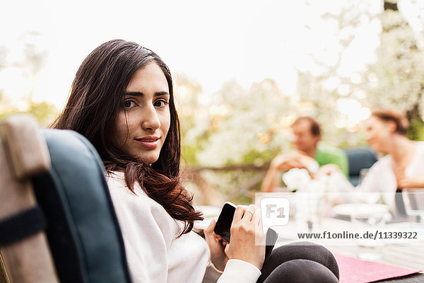 Seitenansicht Porträt eines jungen Mädchens mit Smartphone  während die Eltern im Hintergrund auf dem Hof sitzen.