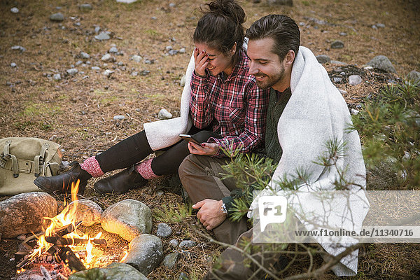High angle view of happy couple wrapped in blanket while using mobile phone by fire pit at campsite