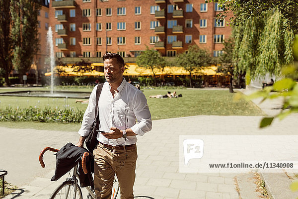 Nachdenklicher Geschäftsmann hält im Sommer Fahrrad und Smartphone auf dem Fußweg.