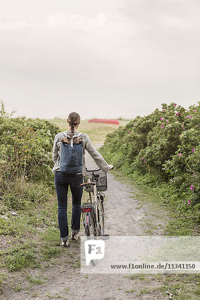Rückansicht einer Frau die einen Rucksack trägt und mit dem Fahrrad auf einem Wanderweg inmitten von Pflanzen wandert.