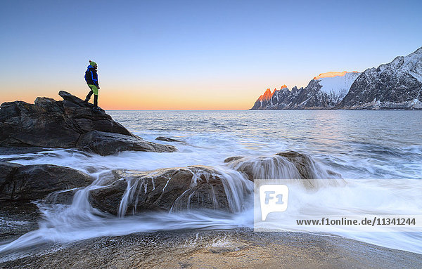 Hiker admires the waves of the icy sea crashing on the rocky cliffs at dawn  Tungeneset  Senja  Troms county  Arctic  Norway  Scandinavia  Europe