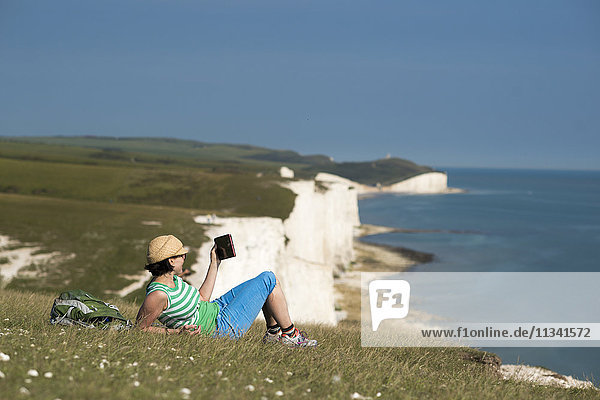 Eine Frau liest auf den Klippen mit Blick auf die Seven Sisters-Küste in der Ferne auf ihrem iPad  South Downs National Park  East Sussex  England  Vereinigtes Königreich  Europa
