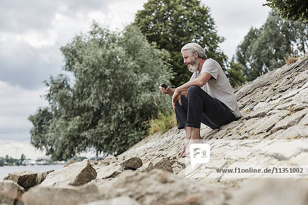 Reifer Mann mit Vollbart sitzt mit Handy am Flussufer