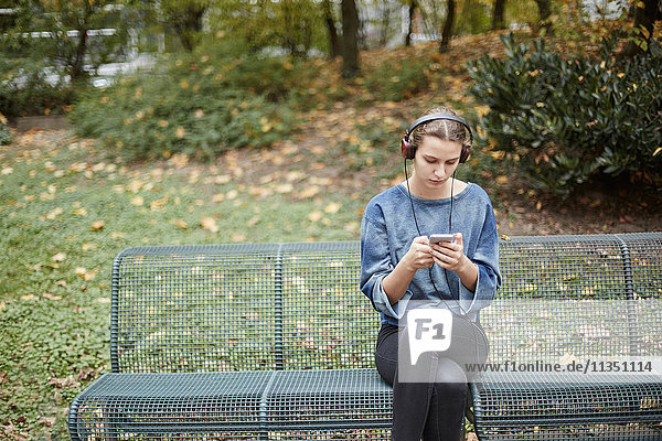 Young woman wearing headphones on a park bench looking at cell phone
