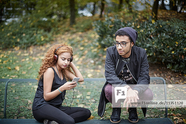 Junges Paar mit Handy und Kopfhörern auf einer Parkbank
