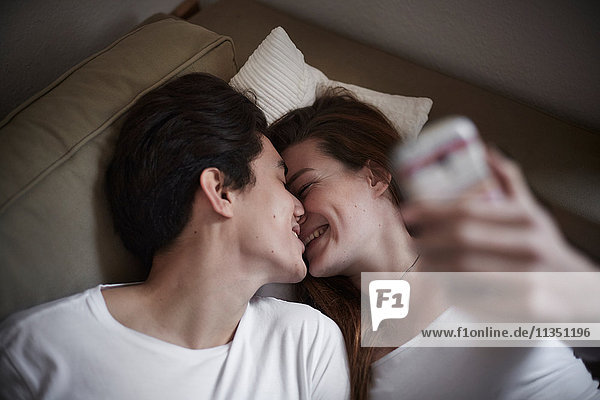 Young couple lying on couch with cell phone kissing