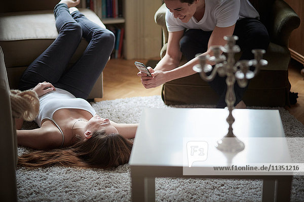 Young man taking cell phone picture of girlfriend lying on carpet on the floor