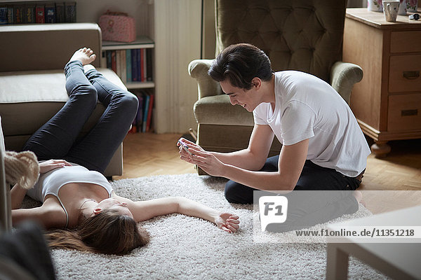 Young man taking cell phone picture of girlfriend lying on carpet on the floor