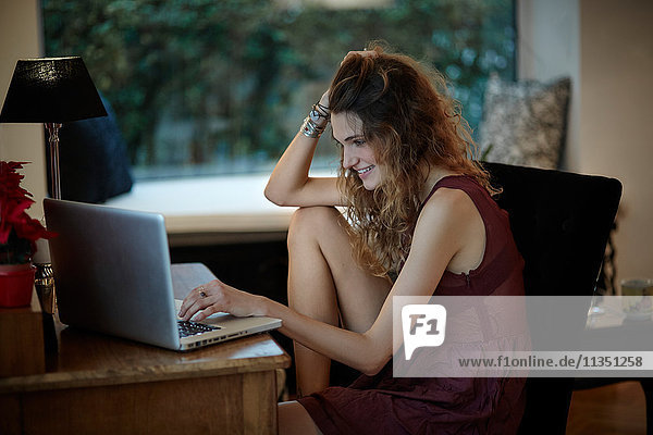 Lächelnde Frau sitzt auf einem Stuhl am Tisch und schaut auf ihren Laptop