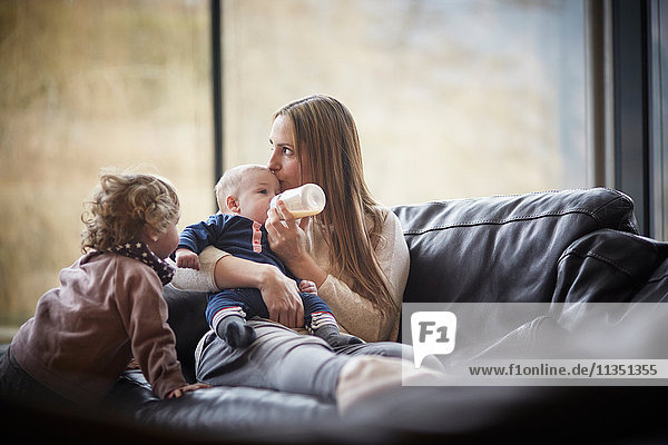 Mutter gibt ihrem Baby das Fläschchen auf der Couch beobachtet von der Tochter