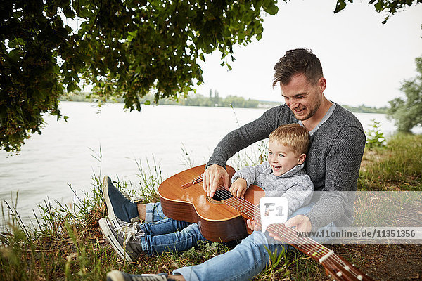 Happy father and son sitting at the riverside playing guitar