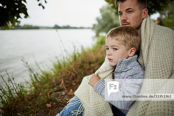 Vater und Sohn sitzen am Flussufer eingewickelt in eine Decke