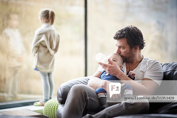 Father bottle feeding baby on couch with daughter at the window