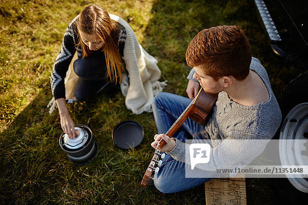 Junges Paar sitzt auf einer Wiese neben einem Auto und einem Campingkocher und macht Musik