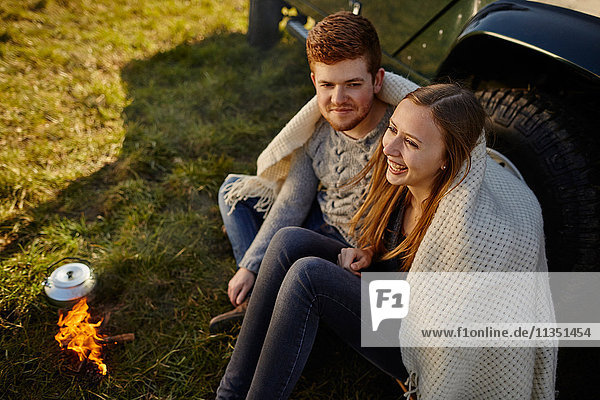 Junges Paar sitzt auf einer Wiese neben einem Auto und Campingkocher