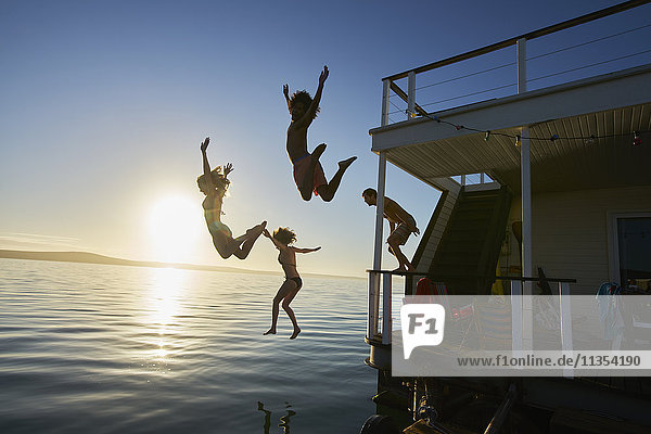 Junge erwachsene Freunde springen vom Sommerhausboot in den Sonnenuntergang.