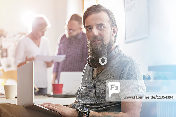 Portrait lächelnder männlicher Designer mit Kopfhörer und Laptop im Büro