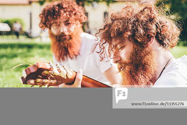 Junge männliche Hipster-Zwillinge mit roten Bärten sitzen im Park und spielen Gitarre