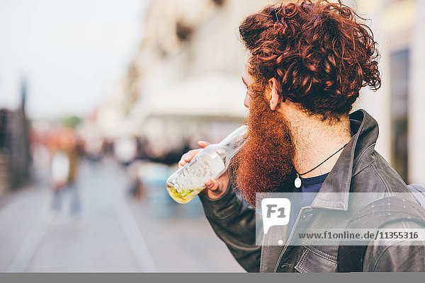 Junger männlicher Hipster mit roten Haaren und Bart  der Flaschenbier trinkt auf der City Street