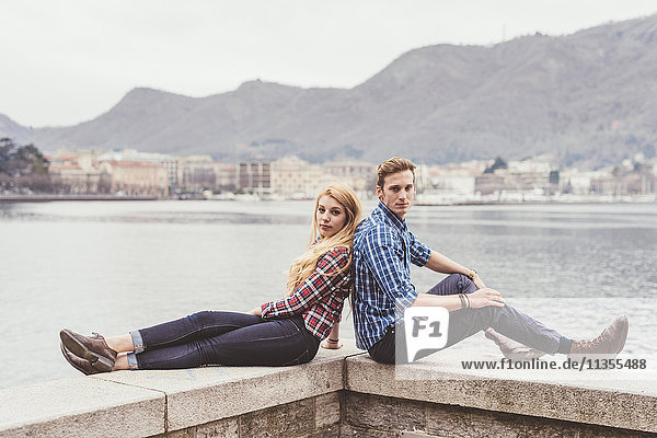Porträt eines jungen Paares  das Rücken an Rücken an der Hafenmauer sitzt  Comer See  Italien