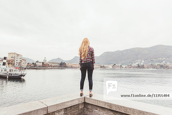 Rückansicht einer jungen Frau  die auf der Hafenmauer steht und nach draußen schaut  Comer See  Italien