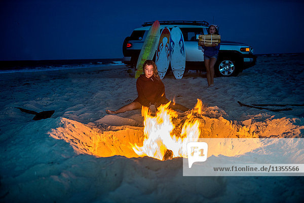 Junges Mädchen sitzt am Lagerfeuer am Strand  ihre Schwester trägt Feuerholz zum Feuer