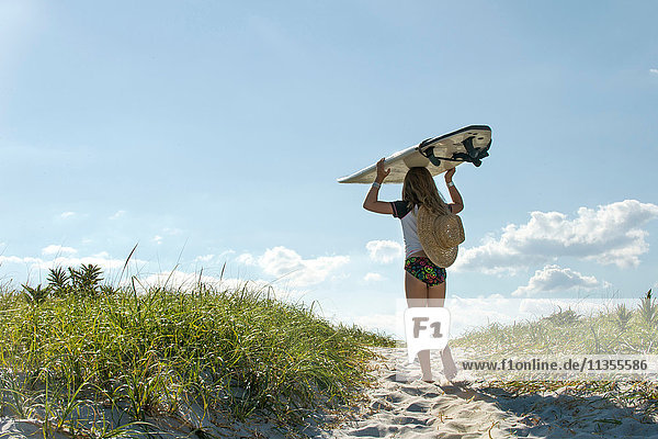 Junges Mädchen geht auf Sanddünen  trägt ein Surfbrett  Rückansicht
