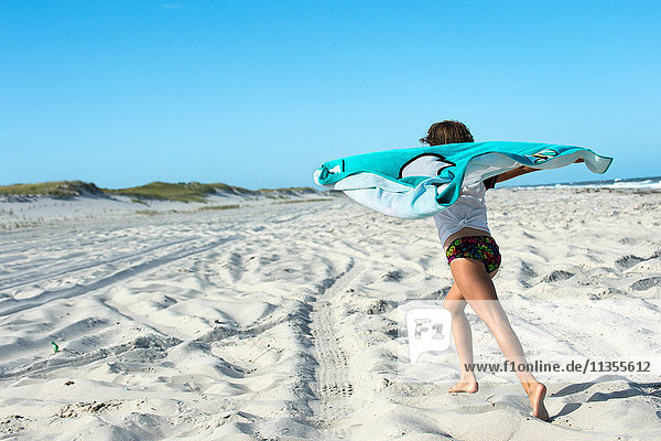 Junges Mädchen läuft auf Sand  hält Strandhandtuch hinter sich  Rückansicht