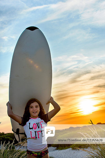 Junges Mädchen am Strand  ein Surfbrett tragend  bei Sonnenuntergang