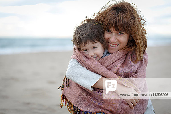 Mutter und Tochter in eine Decke gehüllt umarmen sich am Strand