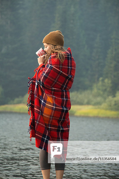 Woman wrapped in tartan blanket drinking coffee by lake  Mount Hood National Forest  Oregon  USA