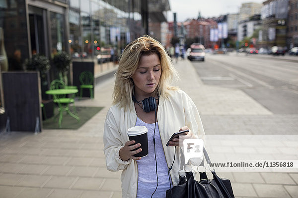 Junge Frau nutzt Smartphone und hält Kaffeetasse auf dem Fußweg in der Stadt