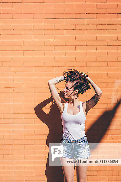 Junge Frau mit Händen in Dreadlock-Haaren vor orangefarbener Wand