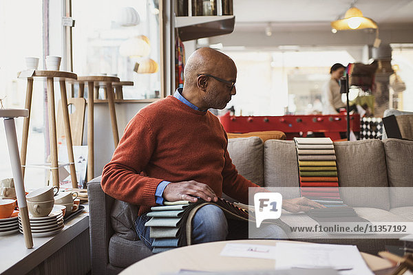 Senior Besitzer arrangiert Stoffmuster  während er auf dem Sofa im Laden sitzt.