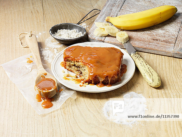 Kochgeschirr und Zutaten für Bananen- und Karamellkuchen,  Holzlöffel mit Karamellsauce,  Mehl,  Banane,  Zucker,  altes Messer und Schneidebrett,  Holztisch