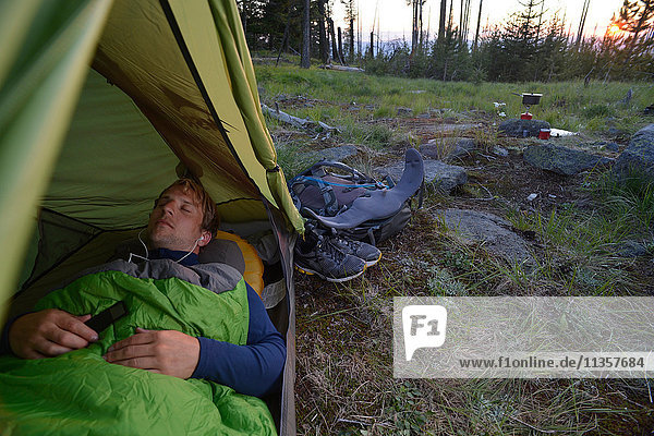 Männlicher Camper im Schlafsack  der in der Abenddämmerung auf dem Midnight Ridge  Colville National Forest  Bundesstaat Washington  USA  Kopfhörer hört