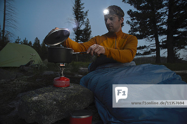 Männlicher Camper im Schlafsack bereitet nachts auf dem Midnight Ridge  Colville National Forest  Bundesstaat Washington  USA  eine Mahlzeit auf dem Campingkocher zu
