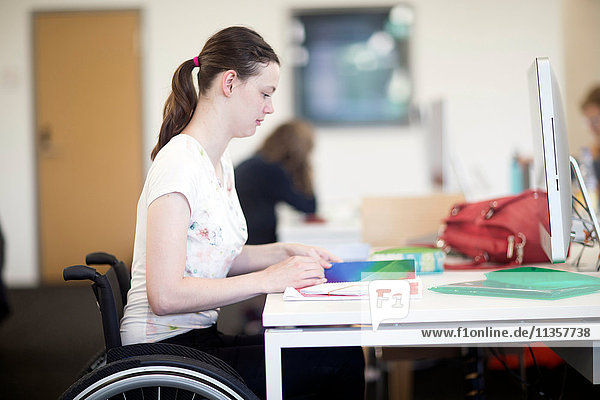 Junge Studentin  die am Schreibtisch Notizen im Rollstuhl liest