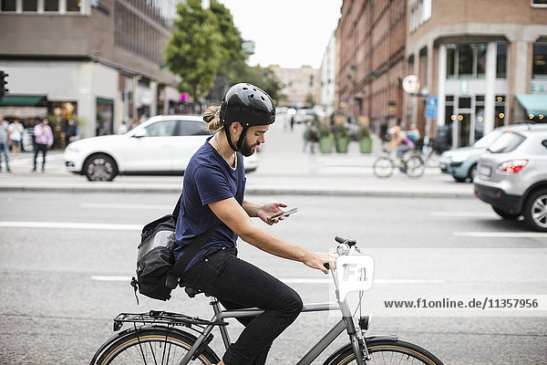 Side view of man using mobile phone while riding bicycle on city street