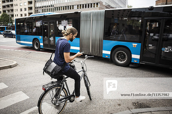 Man with bicycle using mobile phone while standing on city street against articulated bus
