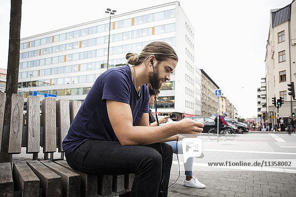 Man listening music through smart phone while sitting beside woman on wooden bench in city