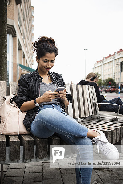 Smiling woman using mobile phone while sitting on wooden bench in city