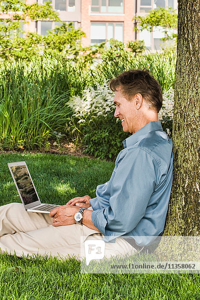 Businessman sitting outdoors  leaning against tree  using laptop