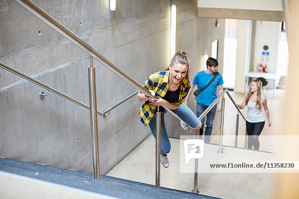 Young female student sliding down stairway handrail at higher education college