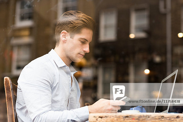 Window view of young businessman reading smartphone texts in cafe