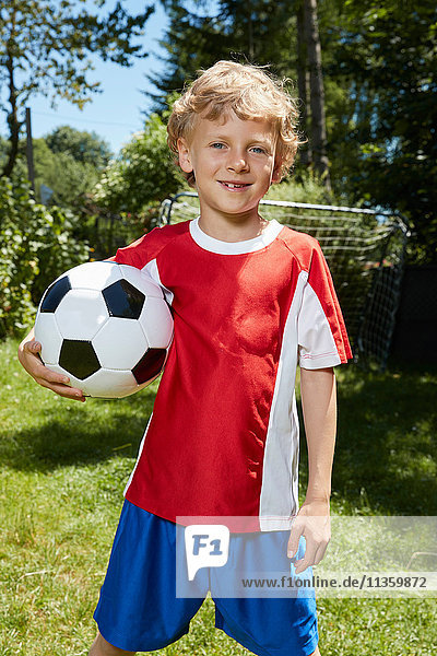Porträt eines Jungen in Fussballuniform  der im Garten einen Fussball hält
