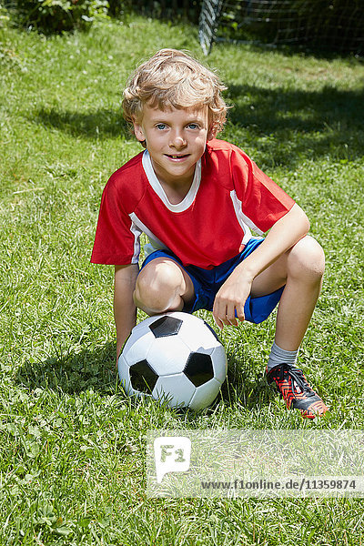 Porträt eines Jungen in Fussballuniform  der mit Fussball im Garten kauert