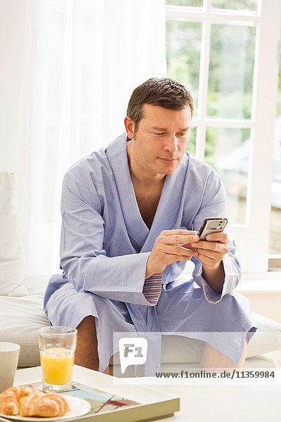 Mature man on sofa reading texts on mobile phone and having breakfast