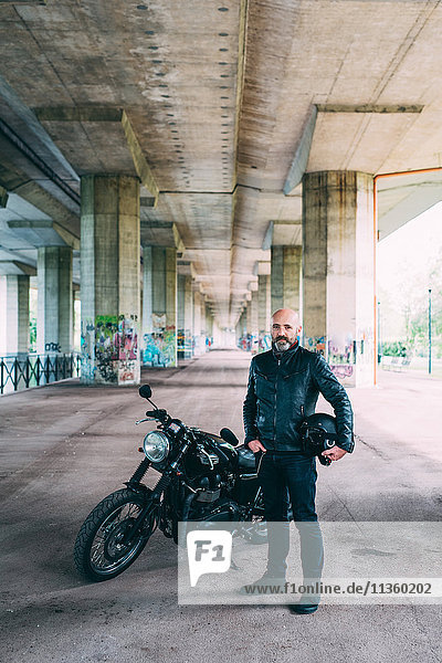 Mature male motorcyclist standing by motorcycle under flyover