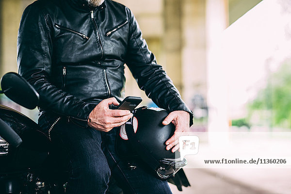 Mid section of mature male motorcyclist sitting on motorcycle texting on smartphone
